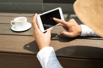 Mockup image of hands holding black mobile under the sun at coffee shop