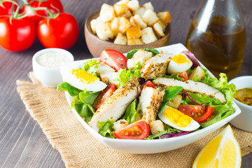 Fresh Caesar salad with delicious chicken breast, ruccola, spinach, cabbage, arugula, egg, parmesan and cherry tomato on wooden background. Oil, salt and pepper. Healthy and diet food concept.