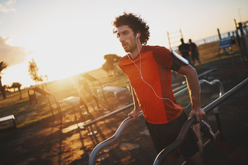 Fit young man listening to music on earphones doing workout on bars in the park on sunny day