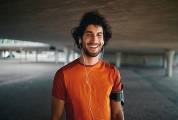 Cheerful portrait of a healthy sporty young man enjoying listening to music on earphones standing on city street - 313594493