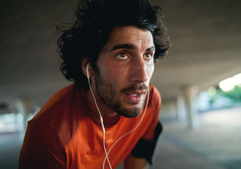 Portrait of a exhausted sweaty serious young male runner with earphone in his ears relaxing after running looking into the distance