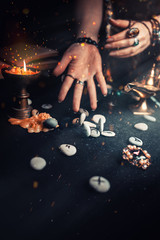 Astrology and esotericism. The female witch's hands read the Runestones, throwing them forward. On a black background lie fortune-telling runes, amulets and a candle. Sparks of fire in the air. Copy