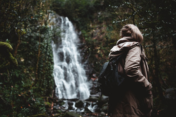 girl with a backpack on her back on a path overgrown with trees and a moss in a dark gloomy forest in the rain and looks at a high powerful waterfall - 313600276