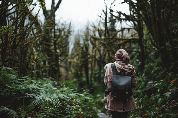 a girl with a rucksack on her back walks along a path overgrown with trees and moss in a dark gloomy forest in the rain - 313600602