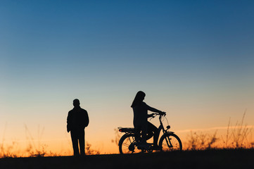 Fototapeta na wymiar Female cyclist on the e-bike or electric bicycle on the sunset background. Silhouette of the woman and man in profile. Active leisure. Travel. Sport.
