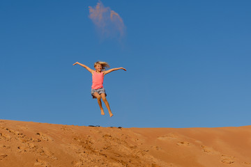 Girl leaping off an orange sand dune with a blue sky background and sand in the air. Fun, excitement, risk taking concept.