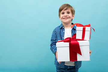 Caucasian boy holds many white boxes with gifts and rejoices, portrait isolated on blue background