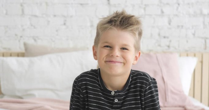 Laughing caucasian child in modern interior. Poratrait of smiling little boy of 5 ages looking at camera sitting in his room on bed. Happy childhood of cute boy. Happiness concept.