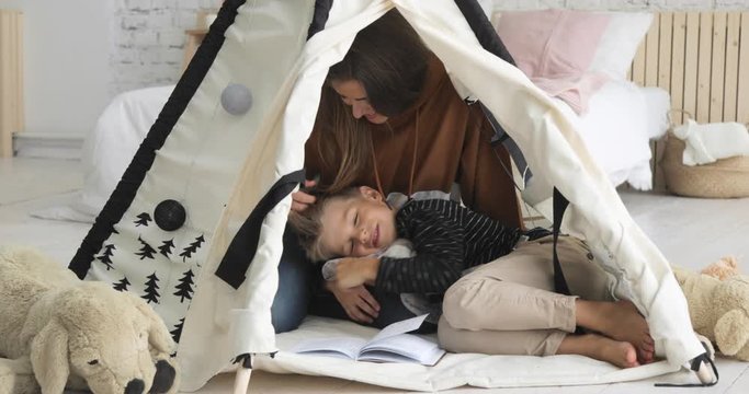 Family at wekend. Mom and son playing together. Mother and son are reading a book in tepee together, boy laying on woman's legs in nursery in modern interior. Happy childhood and motherhood.