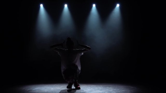 Silhouette guy performs modern hip hop dance on the dark studio with smoke and lighting, twisting and rotating. Slow motion.