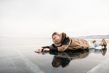 a young girl in a fur coat lies on transparent cracked ice in the middle of the lake, she is happy and smiling - 313611002