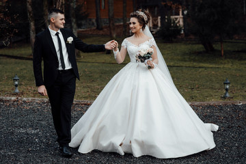 Naklejka na ściany i meble A young and beautiful bride and her husband is standing in park with bouquet of flowers. Amazing smiling wedding couple Beautiful bride and groom on their wedding day. Loving wedding couple outdoor. 