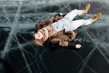 a young girl in a fur coat lies on transparent cracked ice in the middle of the lake, she is happy and smiling - 313611643