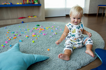 Small cute blond baby sits on a rug and plays with colourful toy bricks around. Toddler in a kinder room. Good for banners