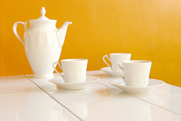 White tea cups and teapot on white tiles table over yellow background..