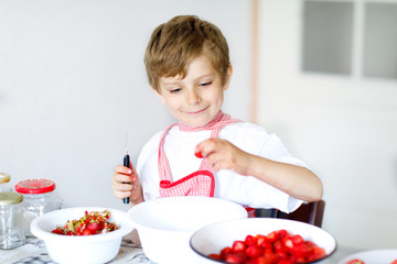 Little blond kid boy helping and making strawberry jam in summer