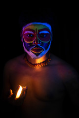 Face of Indian brunette man with his face illuminated and painted by vibrant colors like a tribe standing with candle light in front of a black studio copy space background. Indian hi fashion.