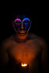 Close up of Indian brunette man with his face illuminated and painted by black and golden like a tribe standing with candle light in front of a black studio copy space background. Indian hi fashion.