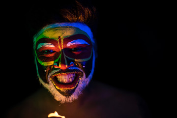 Face of Indian brunette man with his face illuminated and painted by vibrant colors like a tribe standing with candle light in front of a black studio copy space background. Indian hi fashion.