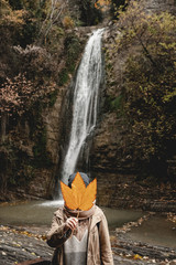 girl holds a large autumn leaf in her hand and covers her face with a waterfall and autumn forest - 313617049
