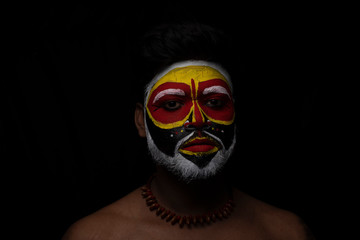 Face of Indian brunette man with his face illuminated and painted by vibrant colors like a tribe standing with candle light in front of a black studio copy space background. Indian hi fashion.