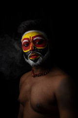 Face of Indian brunette man with his face illuminated and painted by vibrant colors like a tribe standing with candle light in front of a black studio copy space background. Indian hi fashion.