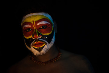 Face of Indian brunette man with his face illuminated and painted by vibrant colors like a tribe standing with candle light in front of a black studio copy space background. Indian hi fashion.