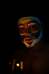 Face of Indian brunette man with his face illuminated and painted by vibrant colors like a tribe standing with candle light in front of a black studio copy space background. Indian hi fashion.