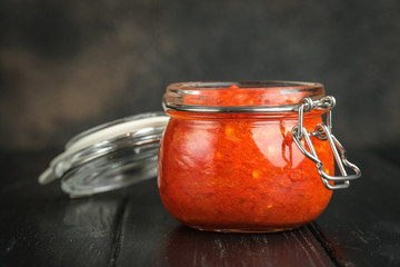 Traditional spicy harissa sauce in a glass jar on a dark background with spices. Maghreb cuisine.