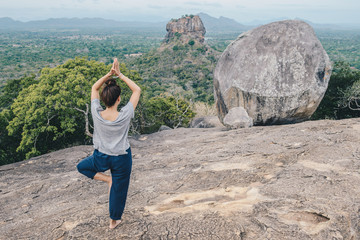 Back view of young woman doing Yoga on top of Pidurangala rock and looking to Sigiriya rock an...