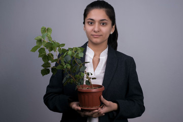 Portrait of a beautiful Indian young business woman in a business suit, holding a green indoor flower in her hand, smiling, rubbing it. On a gray background. Business profit growth concept.