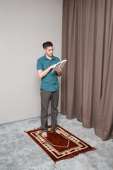 Fototapeta premium A young man prays and recites the muslim Quran on a small portable carpet in the interior. Religion and ritual concept