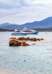 Two luxury boats in the sea. Textured rocks in the foreground, mountains in the background. Vertical landscape. Holidays in Sardinia, Italy.