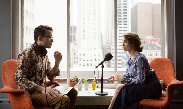 Male journalist interviewing businesswoman while sitting by window in recording studio