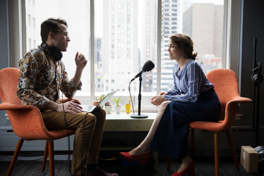 Male journalist interviewing confident businesswoman while sitting by window in recording studio