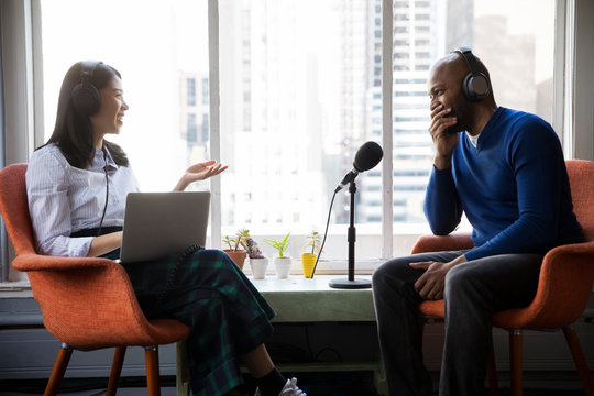 Female journalist interviewing smiling businessman while sitting on chairs by window in recording studio
