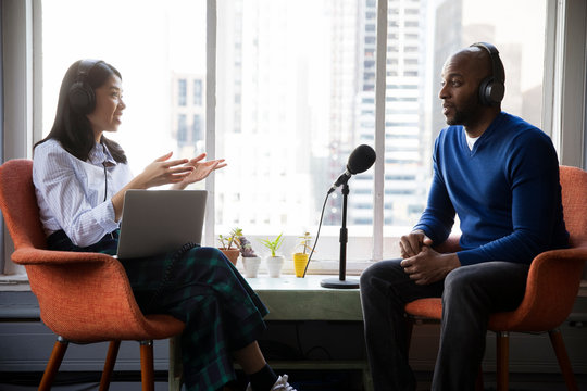 Smiling female journalist interviewing businessman while sitting on chairs by window in recording studio