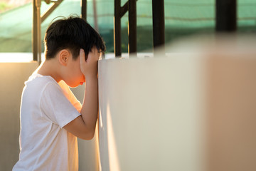 Depressed, frustrated and overwhelmed Asian teenage boy stand alone and lean his head against wall,...