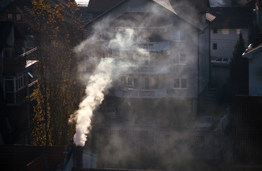 Smoking chimneys at roofs of houses emits smoke, smog at sunrise, pollutants enter atmosphere. Environmental disaster. Harmful emissions and exhaust gases into air. Fog, winter day, heating season.