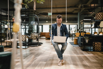Businessman using laptop while sitting on swing in modern office