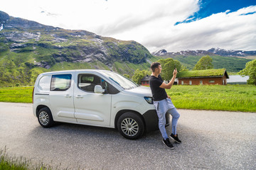 Young handsome tourist man sitting near hired car and use the mobile phone on the norway mountains background. Car hire. Car trip.
