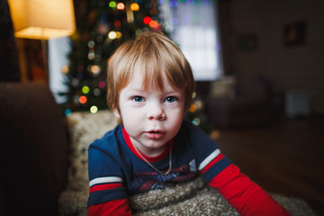 Close-up little blond caucasian boy in the cozy house on the background of a Christmas tree.