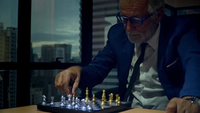 4K Senior business man CEO in blue working suit thinking playing golden and silver chess on chess board game in modern office at night. Management strategy or leadership success concept.