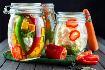 three glass jars of fermented cauliflower, cucumbers, carrots, chili pepper, spices, salt. vegetables on a dark background. using textile green. fermentation is a source of probiotic. vegetarian.