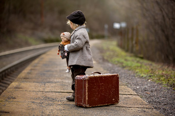 Adorable boy on a railway station, waiting for the train with suitcase and beautiful vintage doll...