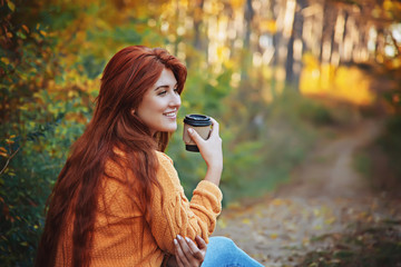 Red-haired smiling woman with a glass of coffee in her hands in the autumn forest.