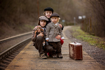 Adorable boys on a railway station, waiting for the train with suitcase and beautiful vintage doll