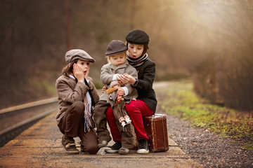 Adorable boys on a railway station, waiting for the train with suitcase and beautiful vintage doll
