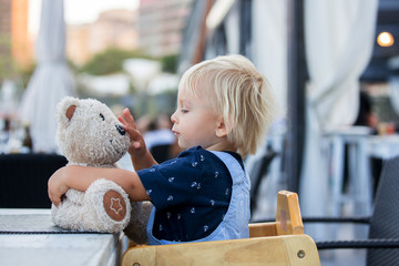 Cute toddler boy, sitting in high chair in a restaurant, playing with teddy bear toy in outdoor beach restaurant