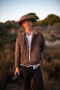 Man with hat, camera, and glasses standing in field at sunset